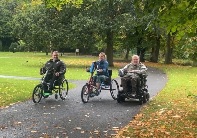 A woman and a man cyling along in hand bikes, with another man in a wheelchair.